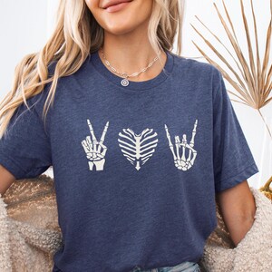 May include: A navy blue t-shirt featuring a white graphic design. The design includes a peace sign, a heart-shaped rib cage, and a rock-and-roll hand gesture. The shirt is made of a soft material.
