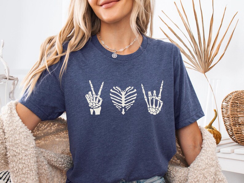 May include: A navy blue t-shirt featuring a white graphic design. The design includes a peace sign, a heart-shaped rib cage, and a rock-and-roll hand gesture. The shirt is made of a soft material.
