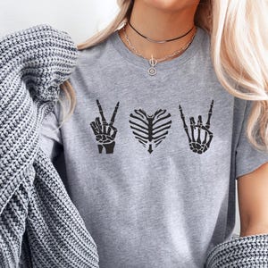 May include: A gray t-shirt featuring three black skeletal designs: a peace sign, a rib cage heart, and a rock-and-roll hand gesture. The shirt is made of a soft material and is suitable for casual wear.