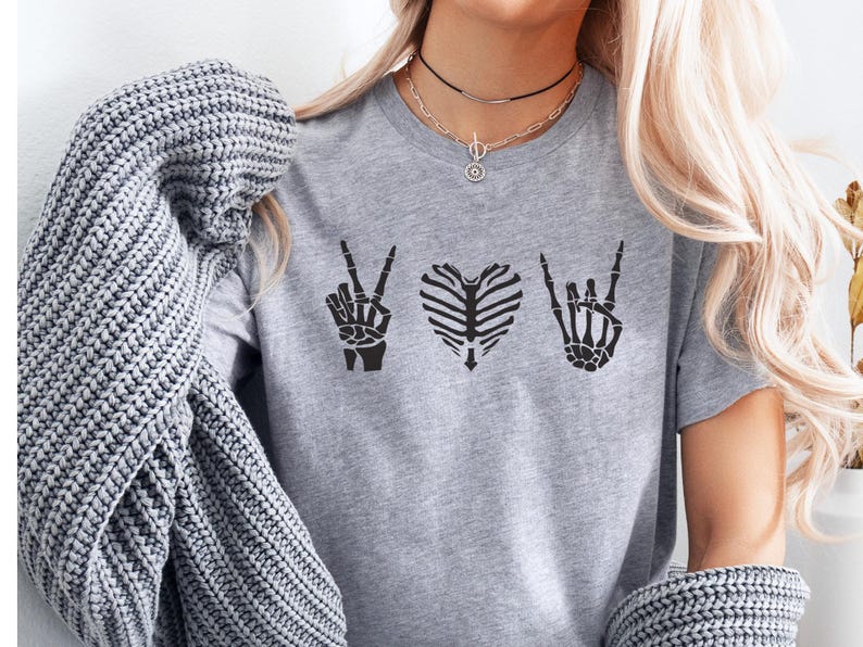 May include: A gray t-shirt featuring three black skeletal designs: a peace sign, a rib cage heart, and a rock-and-roll hand gesture. The shirt is made of a soft material and is suitable for casual wear.