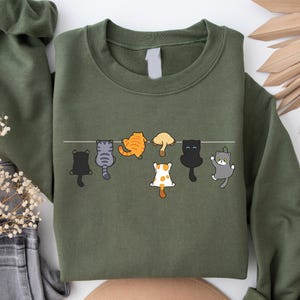 Pode incluir: Camisola verde-oliva com um design extravagante de sete gatos de desenho animado pendurados numa linha branca. Os gatos s&atilde;o de v&aacute;rias cores, incluindo preto, cinza, laranja e branco, com poses divertidas. Uma pe&ccedil;a de vestu&aacute;rio casual e confort&aacute;vel.