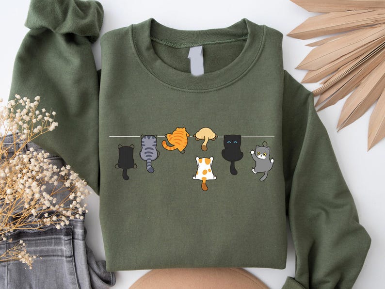 Pode incluir: Camisola verde-oliva com um design extravagante de sete gatos de desenho animado pendurados numa linha branca. Os gatos s&atilde;o de v&aacute;rias cores, incluindo preto, cinza, laranja e branco, com poses divertidas. Uma pe&ccedil;a de vestu&aacute;rio casual e confort&aacute;vel.