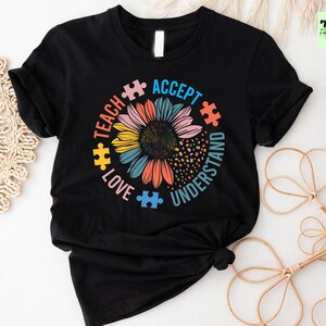 May include: Black t-shirt with a colorful sunflower design and the text "Teach Love Accept Understand" around it. The sunflower is made up of puzzle pieces.