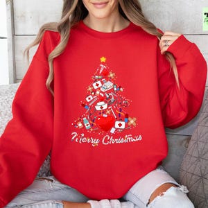 May include: Red sweatshirt with a Christmas tree design made of medical supplies, including a heart, bandages, and syringes. The words "Merry Christmas" are written in white script below the tree. A gold star tops the tree.