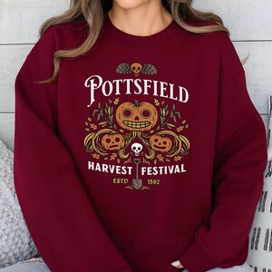 May include: A maroon crewneck sweatshirt featuring a graphic print with the text POTTSFIELD HARVEST FESTIVAL ESTD 1502. The design includes three orange jack-o'-lanterns, green foliage, a small skull, and a shovel centered below the pumpkins.