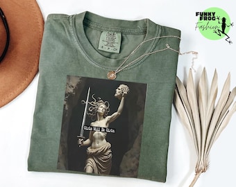 May include: Sage green t-shirt with a graphic of a statue of Medusa holding a sword and a severed head. The graphic includes the text "Girls Will Be Girls." A gold necklace and a dried plant are also visible.