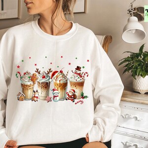 May include: White sweatshirt featuring a festive design of Christmas-themed coffee drinks. Each drink is decorated with whipped cream, candy canes, and holiday characters like gingerbread men, snowmen, and Santa Claus. Red and green accents add to the holiday theme.