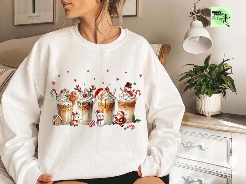 May include: White sweatshirt featuring a festive design of Christmas-themed coffee drinks. Each drink is decorated with whipped cream, candy canes, and holiday characters like gingerbread men, snowmen, and Santa Claus. Red and green accents add to the holiday theme.