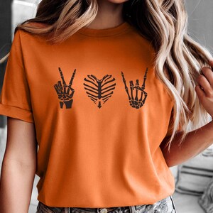 May include: Orange t-shirt with a black skeleton hand peace sign, a rib cage heart, and a skeleton hand rock and roll sign. The shirt is short-sleeved and has a crew neck.