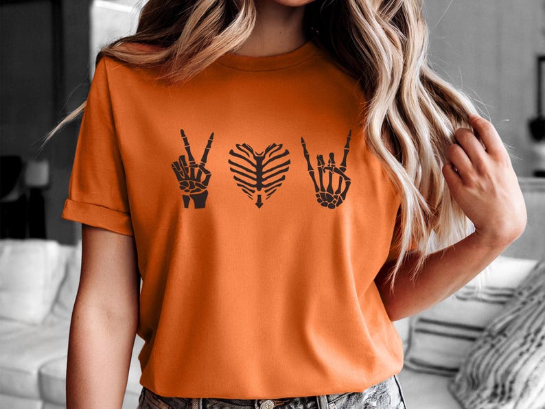 May include: Orange t-shirt with a black skeleton hand peace sign, a rib cage heart, and a skeleton hand rock and roll sign. The shirt is short-sleeved and has a crew neck.