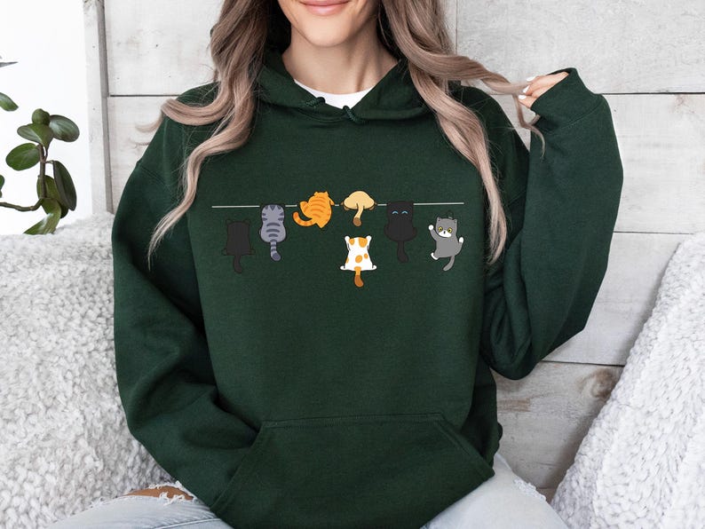 Pode incluir: Moletom verde escuro com um design divertido de gatos de desenho animado pendurados em um varal. Os gatos s&atilde;o de v&aacute;rias cores, incluindo preto, cinza, laranja e branco. O moletom tem capuz com cord&atilde;o e um bolso frontal.