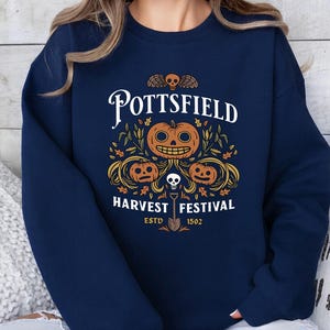 May include: A navy blue crewneck sweatshirt featuring a graphic print with the text POTTSFIELD HARVEST FESTIVAL ESTD 1502. The design includes three orange jack-o'-lanterns, a small skull, autumn leaves, and a shovel centered on the chest.