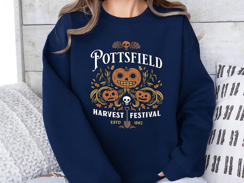 May include: A navy blue crewneck sweatshirt featuring a graphic print with the text POTTSFIELD HARVEST FESTIVAL ESTD 1502. The design includes three orange jack-o'-lanterns, a small skull, autumn leaves, and a shovel centered on the chest.