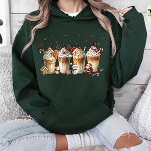 May include: A dark green hoodie featuring a festive design of four decorated coffee drinks. Each drink is topped with whipped cream and festive decorations like gingerbread men, candy canes, and snowmen. The hoodie has a front pocket.