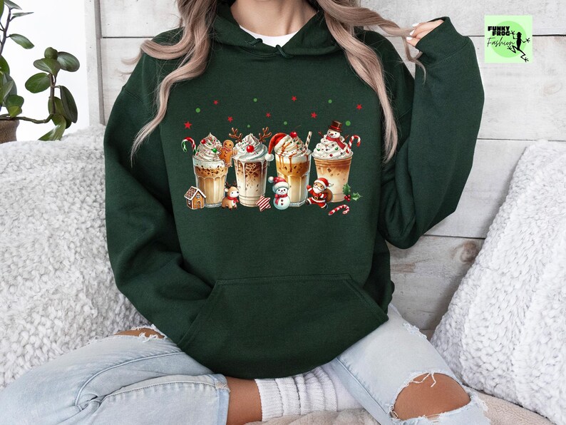 May include: A dark green hoodie featuring a festive design of four decorated coffee drinks. Each drink is topped with whipped cream and festive decorations like gingerbread men, candy canes, and snowmen. The hoodie has a front pocket.
