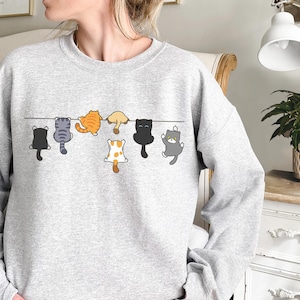 Pode incluir: Um moletom cinza claro com um desenho de desenho animado de gatos pendurados em uma linha preta. Os gatos s&atilde;o de v&aacute;rias cores, incluindo preto, laranja, cinza e branco com manchas marrons. O moletom &eacute; um estilo casual e confort&aacute;vel.