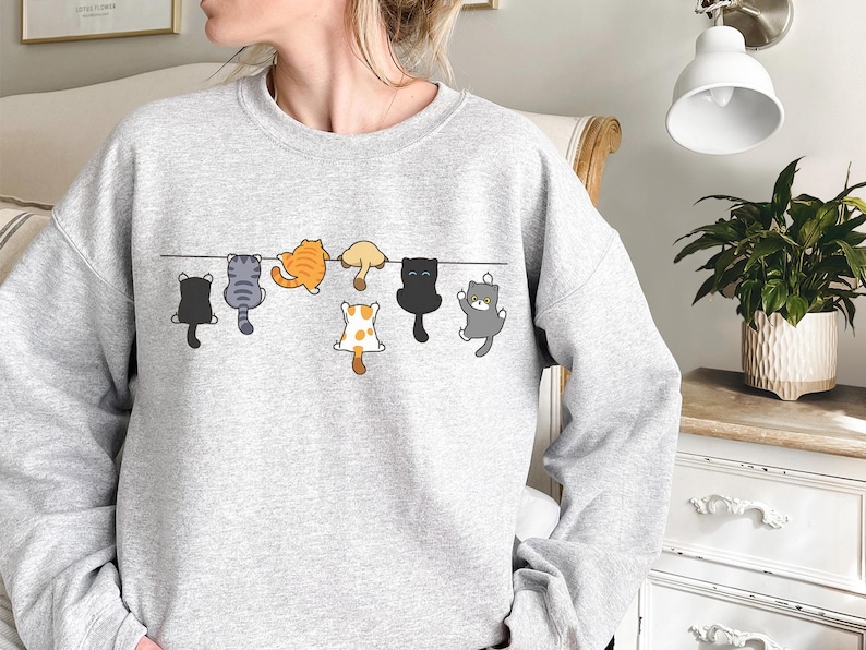 Pode incluir: Um moletom cinza claro com um desenho de desenho animado de gatos pendurados em uma linha preta. Os gatos s&atilde;o de v&aacute;rias cores, incluindo preto, laranja, cinza e branco com manchas marrons. O moletom &eacute; um estilo casual e confort&aacute;vel.