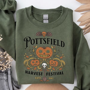 May include: An olive green crewneck sweatshirt featuring a graphic print with the text POTTSFIELD HARVEST FESTIVAL ESTD 1502. The design includes three orange jack-o'-lanterns, a small skull, wheat stalks, autumn leaves, and a shovel.