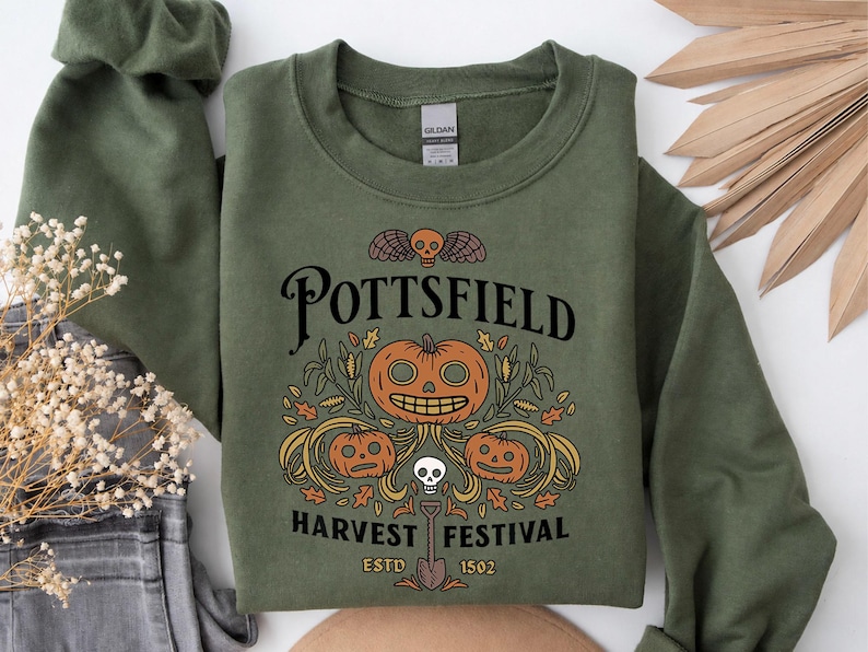 May include: An olive green crewneck sweatshirt featuring a graphic print with the text POTTSFIELD HARVEST FESTIVAL ESTD 1502. The design includes three orange jack-o'-lanterns, a small skull, wheat stalks, autumn leaves, and a shovel.