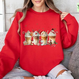 May include: A red sweatshirt featuring a festive graphic of four different holiday-themed coffee drinks. Each drink is decorated with whipped cream, candy canes, and holiday characters like a snowman and Santa Claus. The sweatshirt is a cozy, casual style.