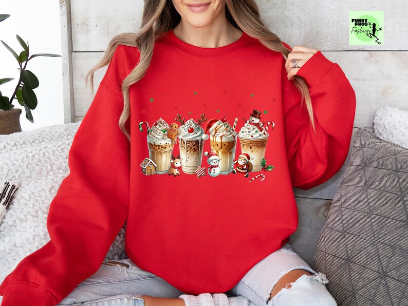May include: A red sweatshirt featuring a festive graphic of four different holiday-themed coffee drinks. Each drink is decorated with whipped cream, candy canes, and holiday characters like a snowman and Santa Claus. The sweatshirt is a cozy, casual style.