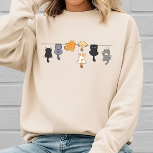 Pode incluir: Camisola bege com um design extravagante de gatos de desenho animado pendurados numa linha. Os gatos s&atilde;o de v&aacute;rias cores, incluindo preto, cinza, laranja e branco, adicionando um toque l&uacute;dico &agrave; roupa casual.
