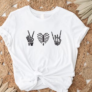 May include: White t-shirt featuring a black graphic design of skeleton hands making the peace sign, a heart made of ribs, and the rock and roll hand gesture. The shirt is knotted at the waist.