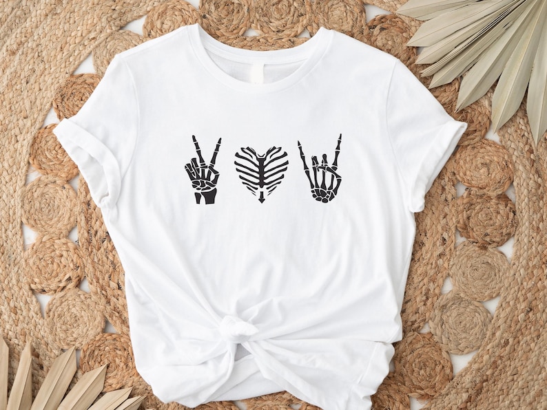 May include: White t-shirt featuring a black graphic design of skeleton hands making the peace sign, a heart made of ribs, and the rock and roll hand gesture. The shirt is knotted at the waist.