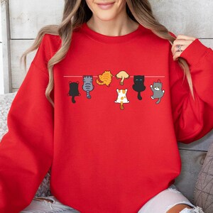 Pode incluir: Um moletom vermelho com um design l&uacute;dico de gatos de desenho animado pendurados em um varal. Os gatos est&atilde;o em v&aacute;rias cores e poses, adicionando um toque divertido &agrave; roupa. O moletom &eacute; um estilo confort&aacute;vel e casual.