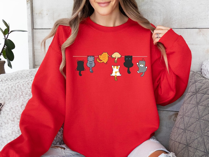 Pode incluir: Um moletom vermelho com um design l&uacute;dico de gatos de desenho animado pendurados em um varal. Os gatos est&atilde;o em v&aacute;rias cores e poses, adicionando um toque divertido &agrave; roupa. O moletom &eacute; um estilo confort&aacute;vel e casual.
