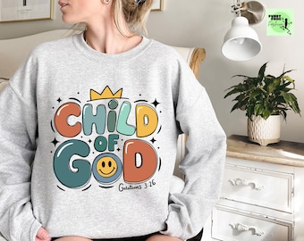 May include: A grey sweatshirt with a colorful graphic design that says "Child of God" with a smiley face and a crown. The text "Galatians 3:26" is also included on the sweatshirt.