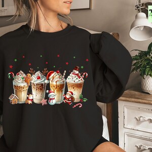 May include: Black sweatshirt featuring a festive design of five holiday-themed coffee drinks with whipped cream, candy canes, and gingerbread men. Red and green star and dot accents complete the holiday theme.