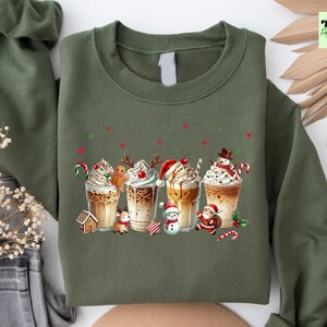 May include: Olive green sweatshirt featuring a festive design of five coffee drinks with whipped cream, candy canes, and holiday characters. The design includes a gingerbread man, reindeer, snowman, and Santa Claus. The sweatshirt has a crew neck and long sleeves.