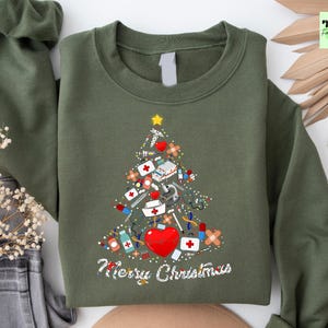 May include: Olive green sweatshirt with a Christmas tree design made of medical supplies, including bandages, syringes, and a red heart. The words "Merry Christmas" are written in white script below the tree. A yellow star tops the tree.