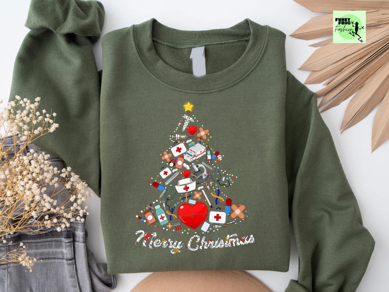 May include: Olive green sweatshirt with a Christmas tree design made of medical supplies, including bandages, syringes, and a red heart. The words "Merry Christmas" are written in white script below the tree. A yellow star tops the tree.