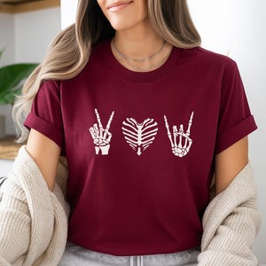 May include: Burgundy t-shirt featuring white skeleton hand gestures: peace, heart, and rock on. The shirt is a crew neck with short sleeves. The design is centered on the chest.