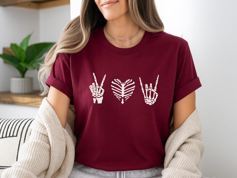 May include: Burgundy t-shirt featuring white skeleton hand gestures: peace, heart, and rock on. The shirt is a crew neck with short sleeves. The design is centered on the chest.