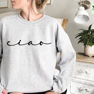 K&ouml;nnte beinhalten: Ein grauer Pullover mit dem Wort "ciao" in schwarzer Kursivschrift.