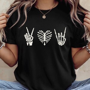 May include: Black t-shirt featuring a white skeleton hand making a peace sign, a rib cage in the shape of a heart, and a skeleton hand making a rock and roll sign. The shirt is a crew neck.