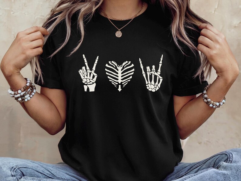May include: Black t-shirt featuring a white skeleton hand making a peace sign, a rib cage in the shape of a heart, and a skeleton hand making a rock and roll sign. The shirt is a crew neck.