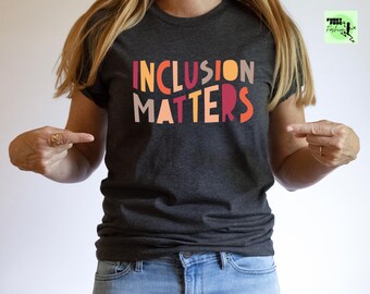 May include: A dark gray t-shirt with the text "INCLUSION MATTERS" in colorful, stylized letters. The letters are in shades of orange, red, pink, and yellow.