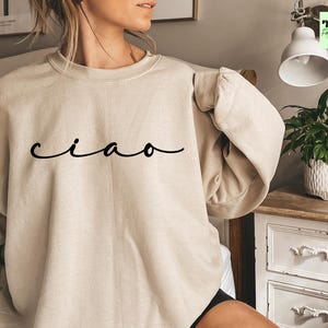 K&ouml;nnte beinhalten: Ein beiger Pullover mit dem Wort "ciao" in schwarzer Kursivschrift.