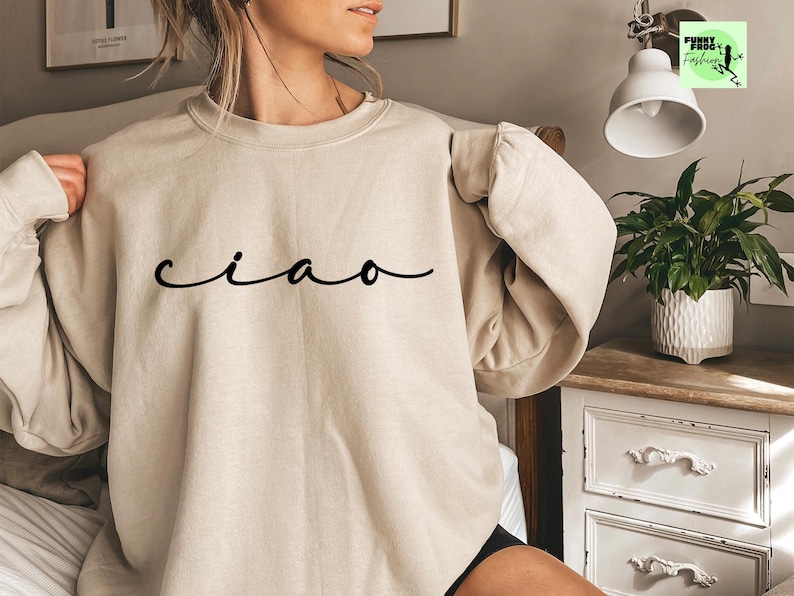 K&ouml;nnte beinhalten: Ein beiger Pullover mit dem Wort "ciao" in schwarzer Kursivschrift.