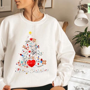 May include: White sweatshirt featuring a Christmas tree design made of medical supplies. The tree includes a red heart, stethoscopes, syringes, bandages, and a yellow star. The words "Merry Christmas" are at the bottom of the tree.
