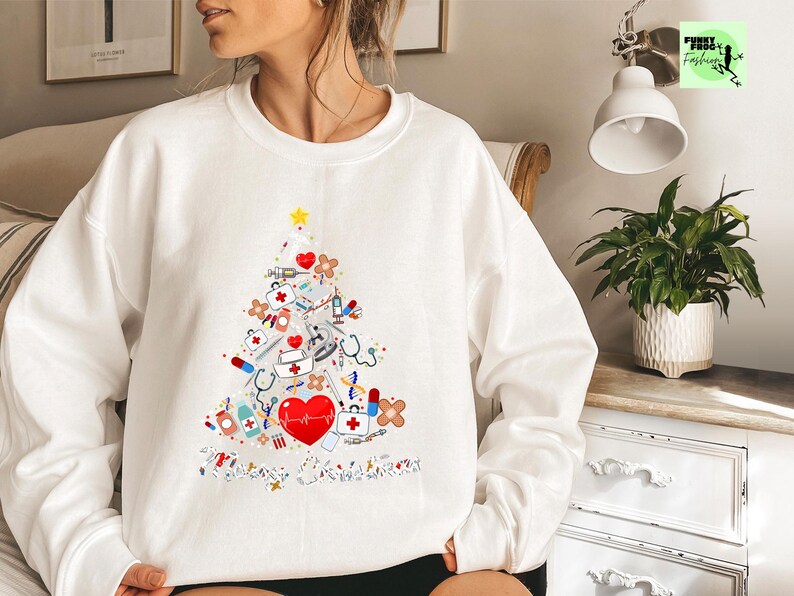 May include: White sweatshirt featuring a Christmas tree design made of medical supplies. The tree includes a red heart, stethoscopes, syringes, bandages, and a yellow star. The words "Merry Christmas" are at the bottom of the tree.