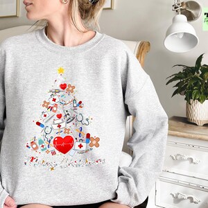 May include: A light gray sweatshirt featuring a Christmas tree design made of medical tools and symbols. The tree includes a red heart, stethoscopes, syringes, and bandages. A yellow star tops the tree, and the design is on the front of the sweatshirt.