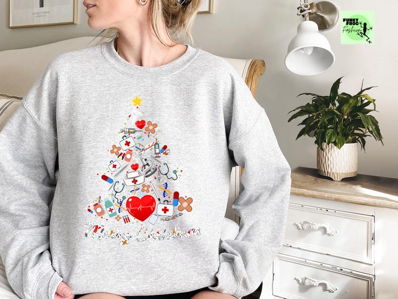 May include: A light gray sweatshirt featuring a Christmas tree design made of medical tools and symbols. The tree includes a red heart, stethoscopes, syringes, and bandages. A yellow star tops the tree, and the design is on the front of the sweatshirt.