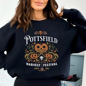 May include: A black crewneck sweatshirt featuring a graphic print with the text POTTSFIELD HARVEST FESTIVAL ESTD 1502. The design includes three orange jack-o'-lanterns, autumn leaves, a small skull with wings, and a shovel centered below.