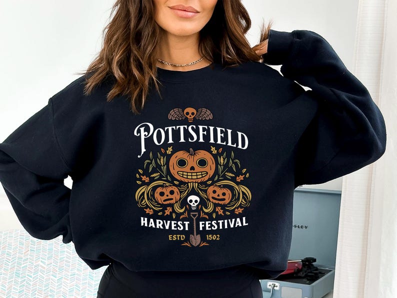 May include: A black crewneck sweatshirt featuring a graphic print with the text POTTSFIELD HARVEST FESTIVAL ESTD 1502. The design includes three orange jack-o'-lanterns, autumn leaves, a small skull with wings, and a shovel centered below.