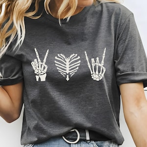 May include: Dark gray t-shirt with a white graphic design featuring a peace sign hand, a rib cage heart, and a rock-on hand gesture. The shirt has short sleeves and a crew neck.